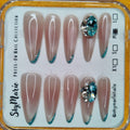 SkyMarie almond press-on nails: nude base, teal glitter French tips, and large teal rhinestone bling.