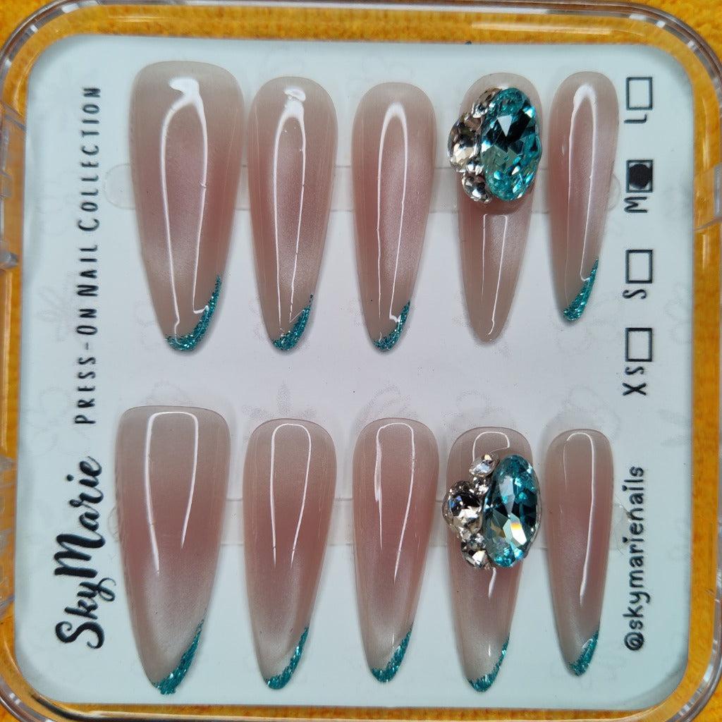 Almond-shaped nude press-on nails with teal glitter French tips and large teal rhinestone accents.