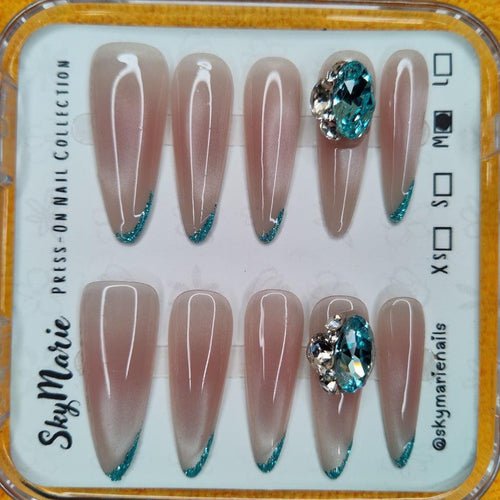 SkyMarie almond press-on nails: nude base, teal glitter French tips, and large teal rhinestone bling.