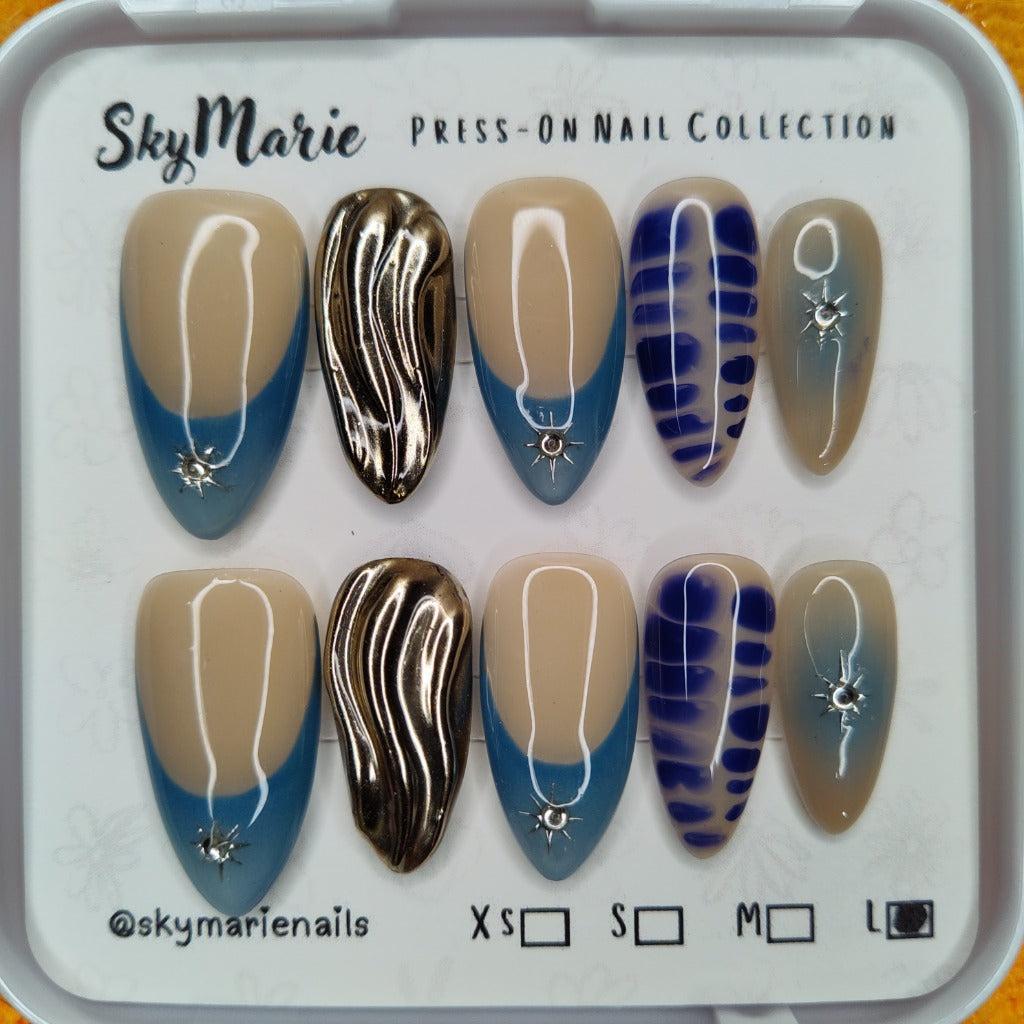 SkyMarie large press-on nails: blue french tips with rhinestones, gold chrome, and abstract blue designs.