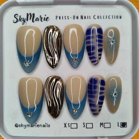 SkyMarie large press-on nails: blue French tips with starburst rhinestones, gold chrome, and abstract blue designs.